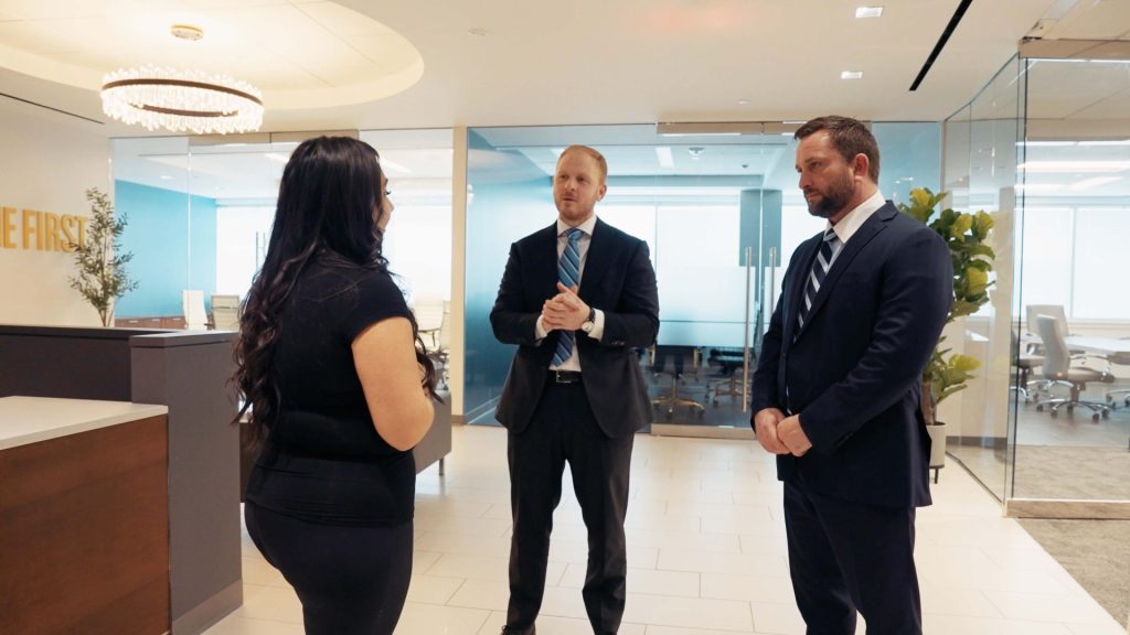 Three people in business attire stand and talk in a modern office lobby with glass walls and a conference room visible in the background.