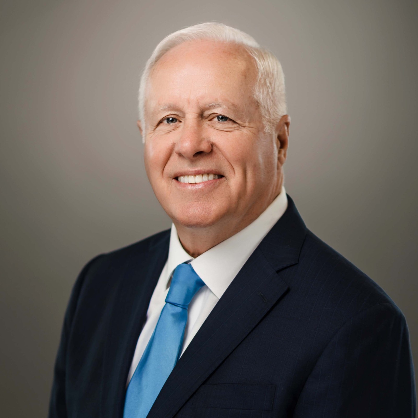 An older man with short white hair, wearing a dark suit, white shirt, and blue tie, poses against a gray background.