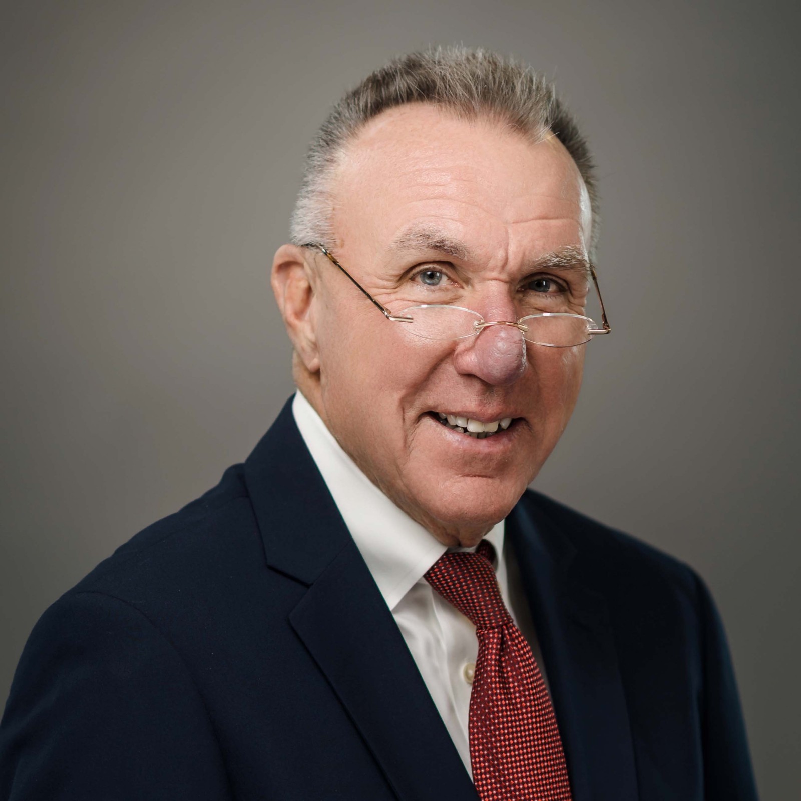 An older man in a dark suit, white shirt, and red tie, wearing glasses, poses against a plain gray background.