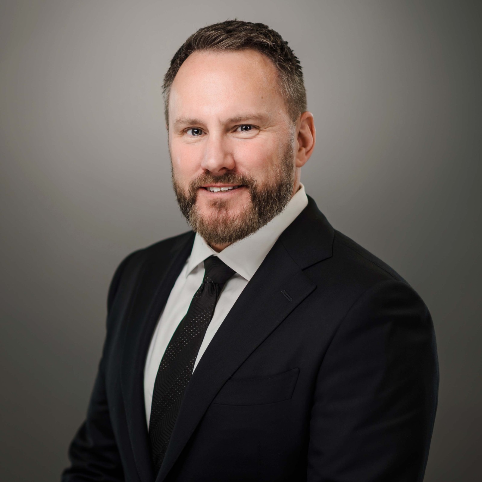 Man in a dark suit, white shirt, and black tie poses for a professional portrait against a plain gray background.